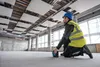 A construction worker is using a laser level in an unfinished room with suspended ceiling panels. Náhľad obrázku produktu