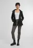 A woman is standing and posing in front of a light gray background, wearing a black jacket over a white top and gray pants, with black boots. Produktbild-Vorschau