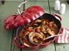 A red Staub cocotte in the shape of a tomato, filled with seafood stew, is placed on a green wooden table. A ladle is scooping some stew from the pot. Two salt and pepper shakers are in the background. Náhľad obrázku produktu
