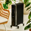 A black oil-filled radiator is shown in a room with plants and a wicker chair. Produktbild-Vorschau