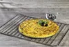 A round, golden-brown dish, possibly a frittata or quiche, sits on a metal cooling rack. It is garnished with fresh green herbs and two black olives. A small glass bowl filled with black olives and a spoon is visible in the background. Produktbild-Vorschau
