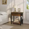 A bedside table with a lamp, picture frame, and charging cables next to a bed. Produktbild-Vorschau