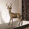 A bronze deer figurine is displayed on a dark wooden shelf against a textured white wall and a stone-patterned background. Produktbild-Vorschau