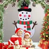 Two children in Christmas hats are sitting in front of a large felt snowman decoration, surrounded by Christmas gifts and a decorated Christmas tree. Náhľad obrázku produktu