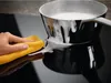 A hand is cleaning a black induction cooktop with a yellow cloth, and a pot with foamy liquid is on the cooktop. Voir les images du produit
