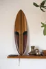 A decorative wooden surfboard hangs on a wall above a shelf with decorative items. Produktbild-Vorschau