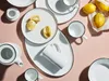 A collection of white ceramic dishes and a teapot with black trim, along with lemons, are arranged on a pink surface. Produktbild-Vorschau