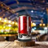 A red beverage dispenser sits on a wooden surface in front of a blurred background of lights. Náhled obrázku produktu
