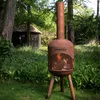 A rusty metal fire pit with a chimney stands in a grassy area with trees and a small wooden structure in the background. Anteprima dell’immagine prodotto