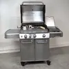 A stainless steel gas grill with its lid open, showing the cooking grates and some accessories. Podgląd zdjęcia produktu