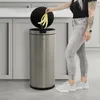 A person is throwing banana peels into a silver sensor trash can in a kitchen. Produktbild-Vorschau