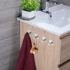 A bathroom vanity with a sink, a plant, and four adhesive hooks on the cabinet door, one of which is holding a maroon towel and a wooden-handled brush. Produktbild-Vorschau