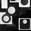 A flat lay arrangement of black and white plates and saucers, with geometric black shapes and a piece of dark material. Náhled obrázku produktu