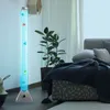 A tall, clear tube filled with blue water and small decorative fish, standing on a wooden floor next to a bed and a plant. Produktbild-Vorschau