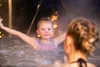 A young girl with her arms outstretched is in a hot tub with another person's back visible in the foreground. Steam rises around them, and blurred lights are visible in the background. Náhled obrázku produktu