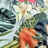 A close-up view of a fabric with a tropical floral pattern, showing hibiscus flowers and large green leaves. Produktbild-Vorschau