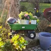 A green garden cart filled with plants and soil, set in a garden with flowers and a tree. Náhled obrázku produktu