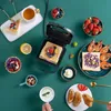 A breakfast spread featuring a sandwich maker with a sandwich inside, along with various fruits, shrimp, and other food items arranged on a green surface. Náhled obrázku produktu