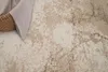 A close-up view of a rug with a distressed pattern in beige and brown tones. A corner of a woven basket and a light-colored fabric are visible in the top left. Produktbild-Vorschau