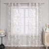A pair of sheer curtains with a floral pattern hangs in front of a window, with a small table and lamp to the left and a cabinet with decorative vases to the right. Produktbild-Vorschau
