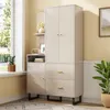 A modern, beige wardrobe with gold accents is placed in a room with a window and a plant. The wardrobe has doors and drawers, and a small shelf unit with decorative items. Produktbild-Vorschau