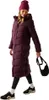 A woman wearing a long burgundy puffer coat and a matching beanie, holding a black bag. Voir les images du produit