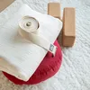 Yoga props including a rolled-up yoga strap, a white folded blanket, a red bolster, and two cork yoga blocks are arranged on a textured white rug. Produktbild-Vorschau
