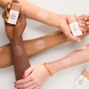 Multiple arms with different skin tones are shown, with swatches of a product applied to them. Two small bottles of the product are also visible. Voir les images du produit