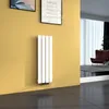 A vertical radiator is mounted on a yellow wall in a modern living room with a brown armchair and sheer curtains in the background. Produktbild-Vorschau