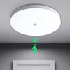 A white circular LED ceiling light with a motion sensor is mounted on a gray ceiling. A green graphic of a person walking with motion waves emanates from below the light, indicating its motion-sensing feature. Náhľad obrázku produktu