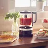 A KitchenAid food processor filled with chopped tomatoes sits on a wooden countertop next to a vase of parsley and a plate of food. Voir les images du produit