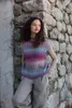 A woman stands leaning against a stone wall, wearing a colorful knitted vest over a beige turtleneck and gray pants. Produktbild-Vorschau
