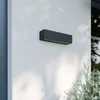 A dark gray rectangular LED wall light is mounted on a white wall, with shadows of leaves cast on the wall and a plant in the foreground. Produktbild-Vorschau