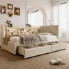 A beige daybed with storage drawers and a bookshelf headboard is shown in a well-lit room with sheer curtains and decorative items on the shelves. Náhled obrázku produktu