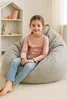 A young girl is sitting on a large, light gray beanbag chair in a room with a white bookshelf in the background. Produktbild-Vorschau