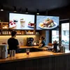 Two people working behind a counter in a cafe, with screens displaying food and drinks above them. Anteprima dell’immagine prodotto