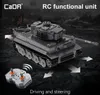 A grey toy tank with a remote control in front of it, set against a dark, blurred background suggesting a battlefield. Náhľad obrázku produktu