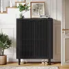 A black cabinet with slatted doors is placed in a hallway next to a staircase, with a plant and a framed picture on top. Produktbild-Vorschau