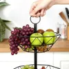 A hand holding a two-tiered black metal fruit basket filled with grapes, apples, and kiwis. Produktbild-Vorschau