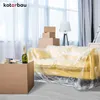 A living room being prepared for moving or renovation, with a yellow sofa covered in plastic sheeting, moving boxes, and a bottle of champagne. Náhľad obrázku produktu