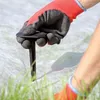 A gloved hand is shown pushing a black plastic anchor into a white landscape fabric laid over grass. Náhled obrázku produktu