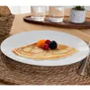 A close-up of a dessert plate with a crepe topped with berries and apricot slices, set on a woven placemat. Produktbild-Vorschau
