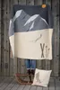A person is standing on a sled, holding up a blanket with a mountain and ski design. A pillow with a skier design is on the ground next to the sled. Produktbild-Vorschau
