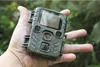 A hand holds a small black and gray trail camera in an outdoor, leafy environment. Produktbild-Vorschau