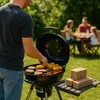 A man is grilling burgers and hot dogs in a backyard while a family sits at a table in the background. Produktbild-Vorschau