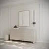 A modern dresser with a textured front and round legs, placed in a minimalist room with wall paneling and abstract art. Voir les images du produit