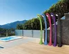A row of colorful outdoor showers are set up next to a hot tub on a wooden deck, with a scenic mountain view in the background. Produktbild-Vorschau