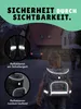 A child wearing a backpack walks away from the camera on a street at dusk, with houses and a car in the background. The backpack and shoulder straps have reflective elements that are illuminated. Produktbild-Vorschau