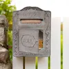 A rustic, vintage-style metal mailbox mounted on a white picket fence, with a stone pillar to the left and greenery in the background. Podgląd zdjęcia produktu