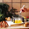 A hand is shown using a whisk to beat eggs in a glass bowl, surrounded by fresh vegetables like carrots and lettuce, on a kitchen counter. Produktbild-Vorschau